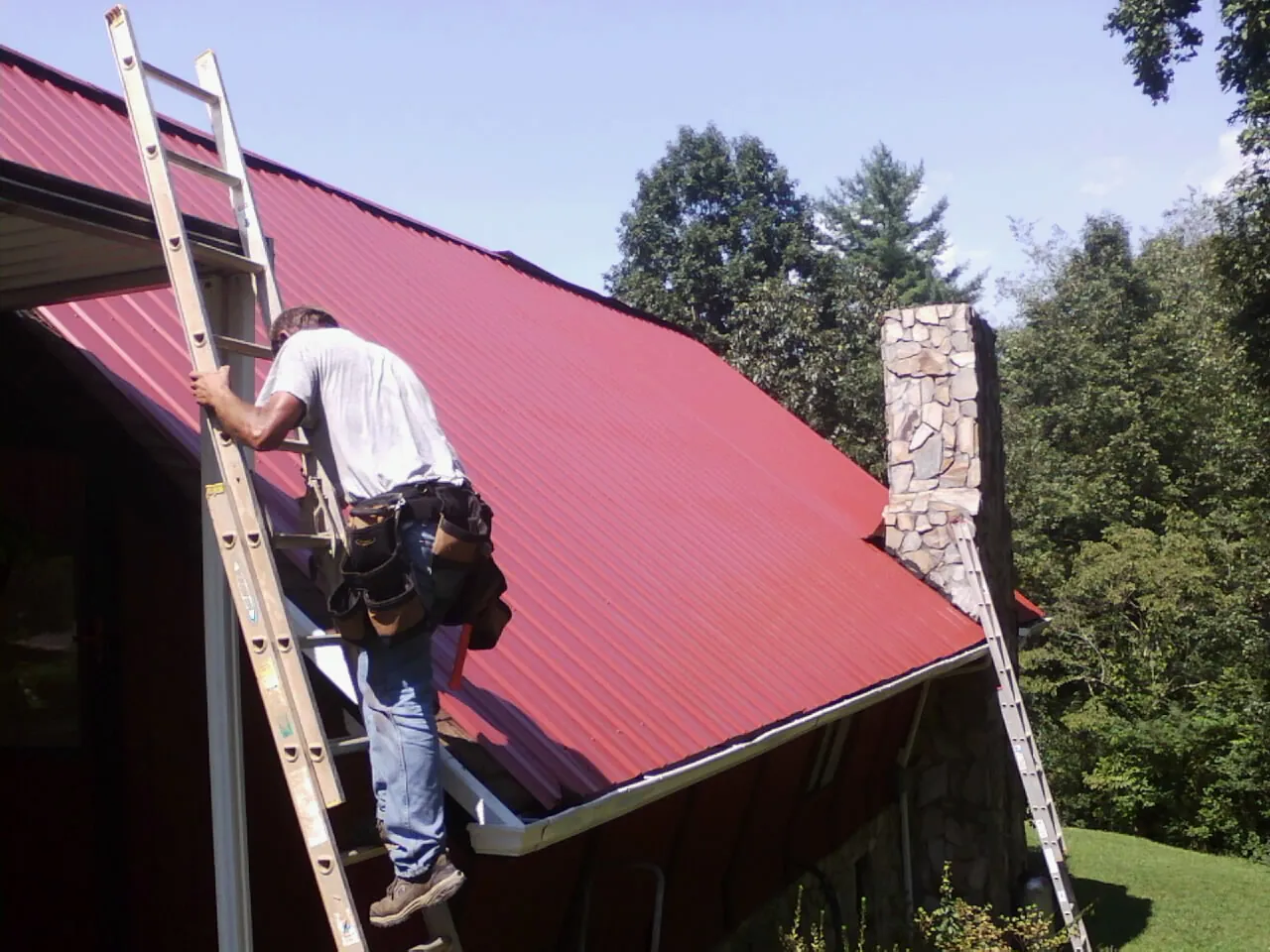 Roofer on a ladder beside a red metal roof and stone chimney — Soffit & Fascia Repair by Roofing Brothers in Healdsburg